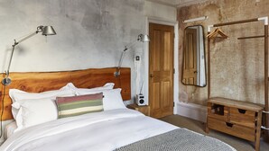 A bedroom with a wooden headboard, a bed with white bedding, two bedside lamps, a radio, a wooden bench, and a mirror on the wall. - The Culpeper (London)