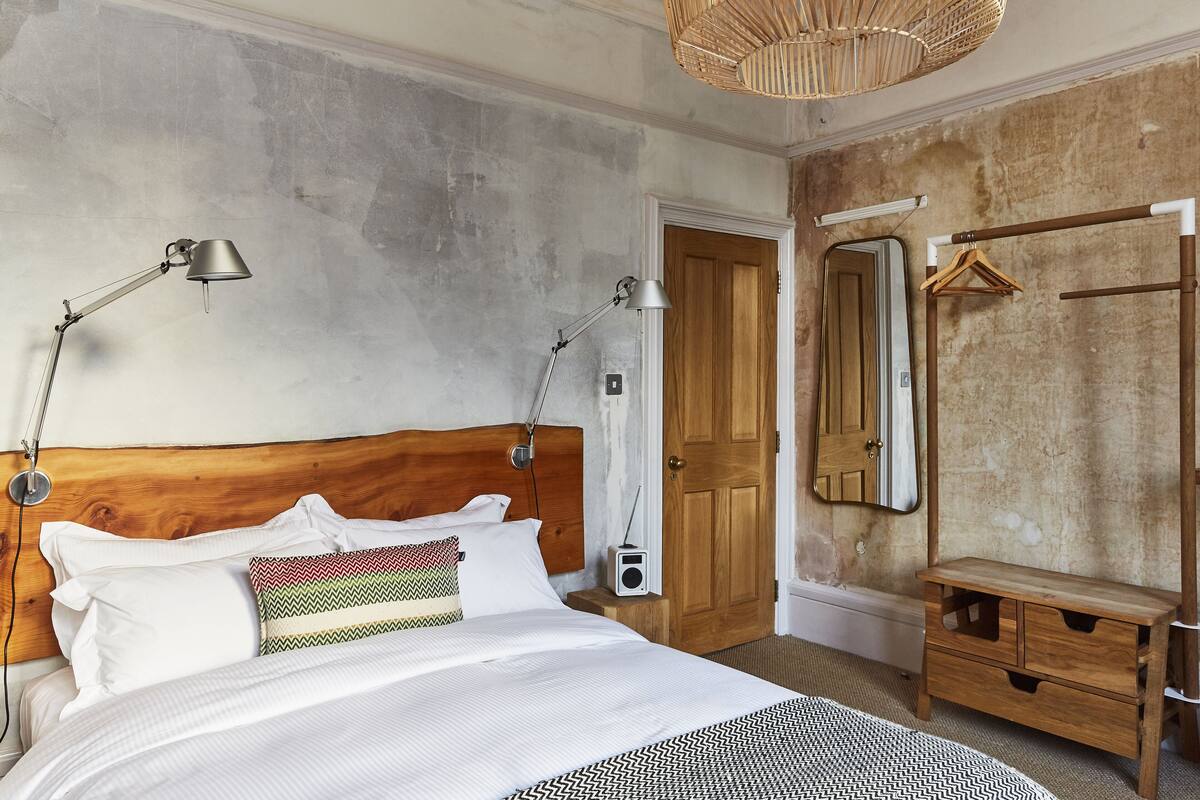Een slaapkamer met een houten hoofdeinde, een bed met wit beddengoed, twee bedlampjes, een radio, een houten bankje en een spiegel aan de muur.