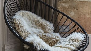 A black wireframe chair with a fluffy white throw blanket, positioned against a wall with a window and curtains. - The Culpeper (London)