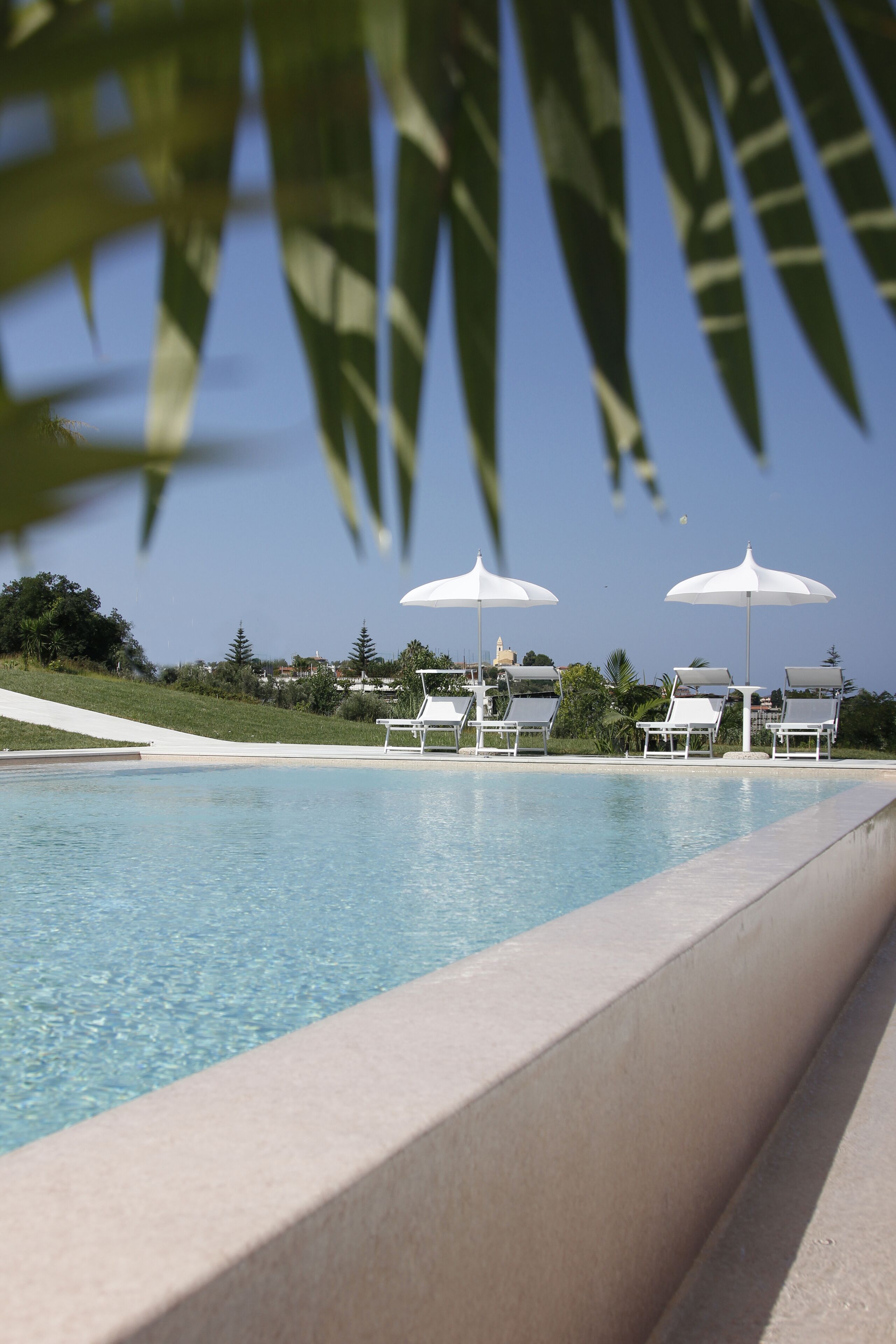Una piscina con sillas de descanso y sombrillas, rodeada de vegetación y un cielo despejado.