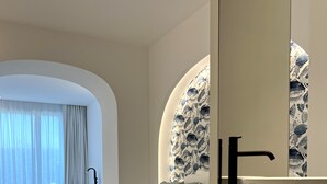A modern bedroom with a large bed, a built-in vanity with a mirror, and a view of the outdoors through a window with sheer curtains. - Il Borghetto Resort & Spa (Tropea)