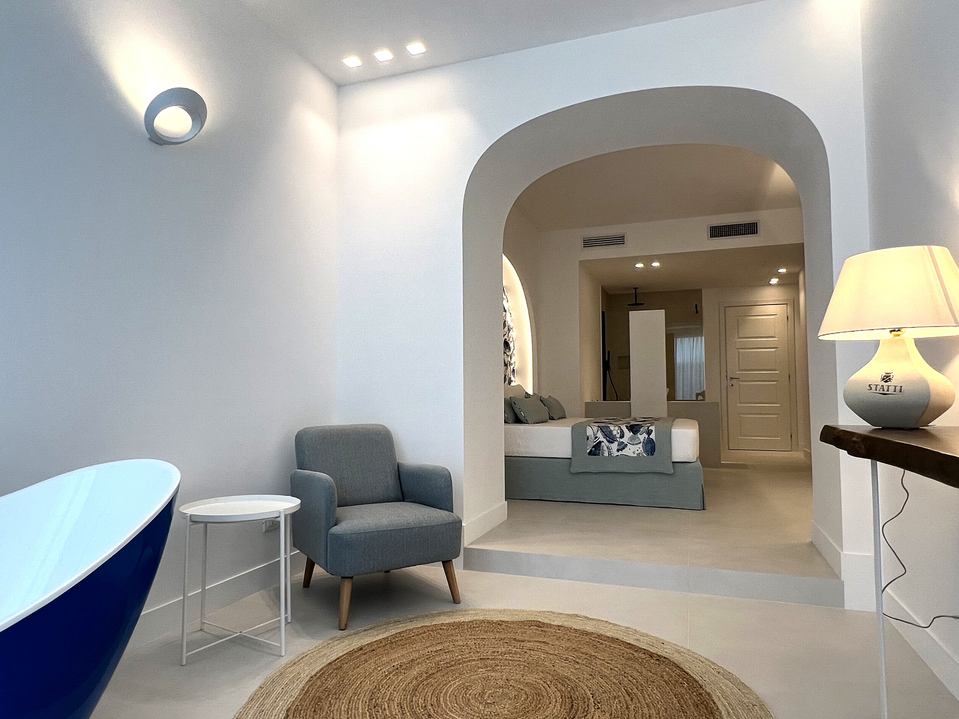 A modern hotel room with a blue bathtub, a grey armchair, a small round table, and a lamp on a wooden side table.