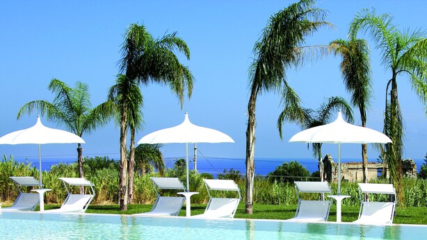 A pool area with white lounge chairs and umbrellas, palm trees, and a view of the ocean. - Il Borghetto Resort & Spa (Tropea)