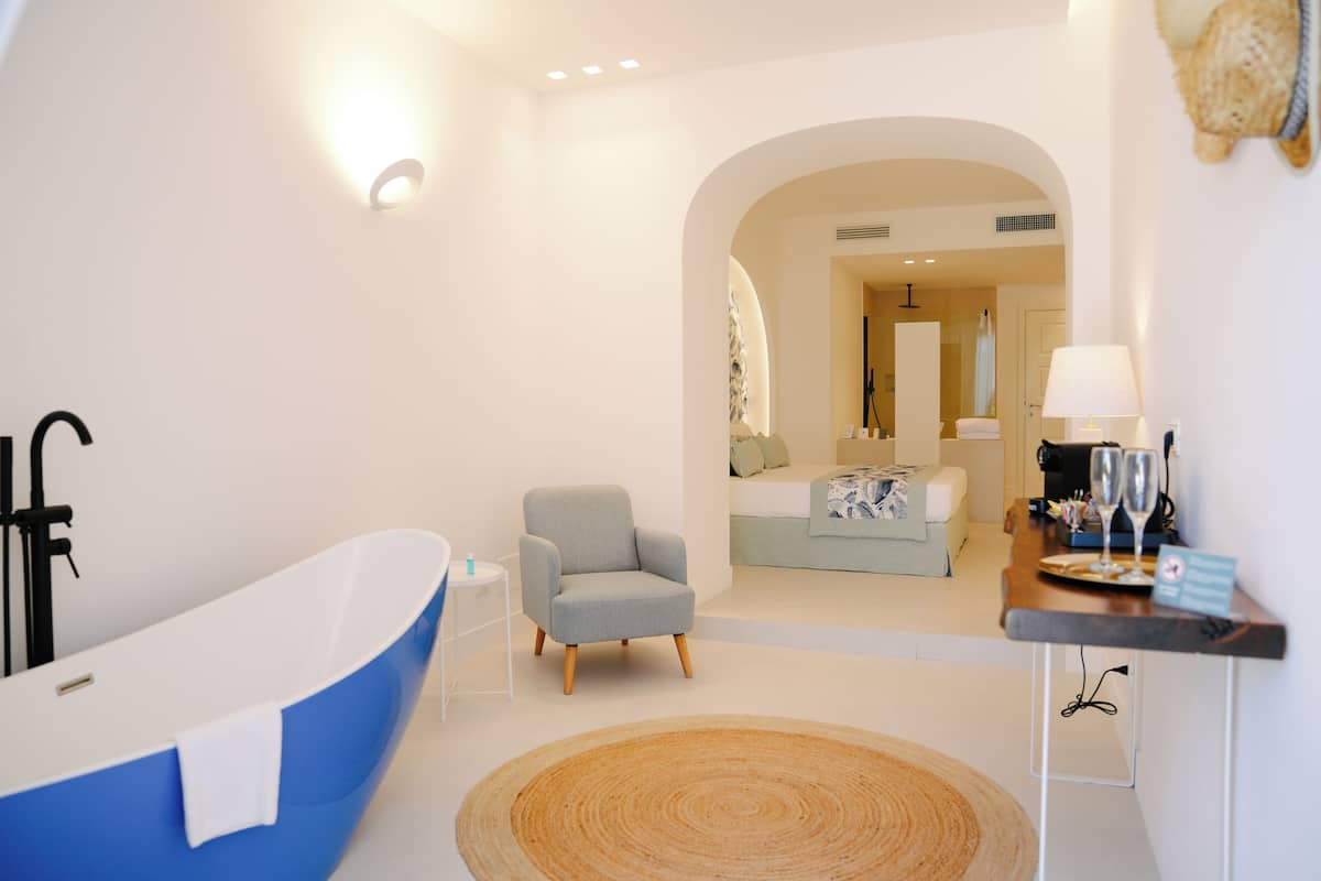 A modern bathroom with a freestanding bathtub, a small armchair, and a bedside table with a lamp and glasses.