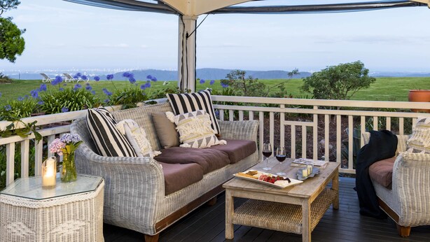 A covered outdoor seating area with wicker furniture, a glass table, and a view of a green landscape. - Rosewood Estate (Beechmont)
