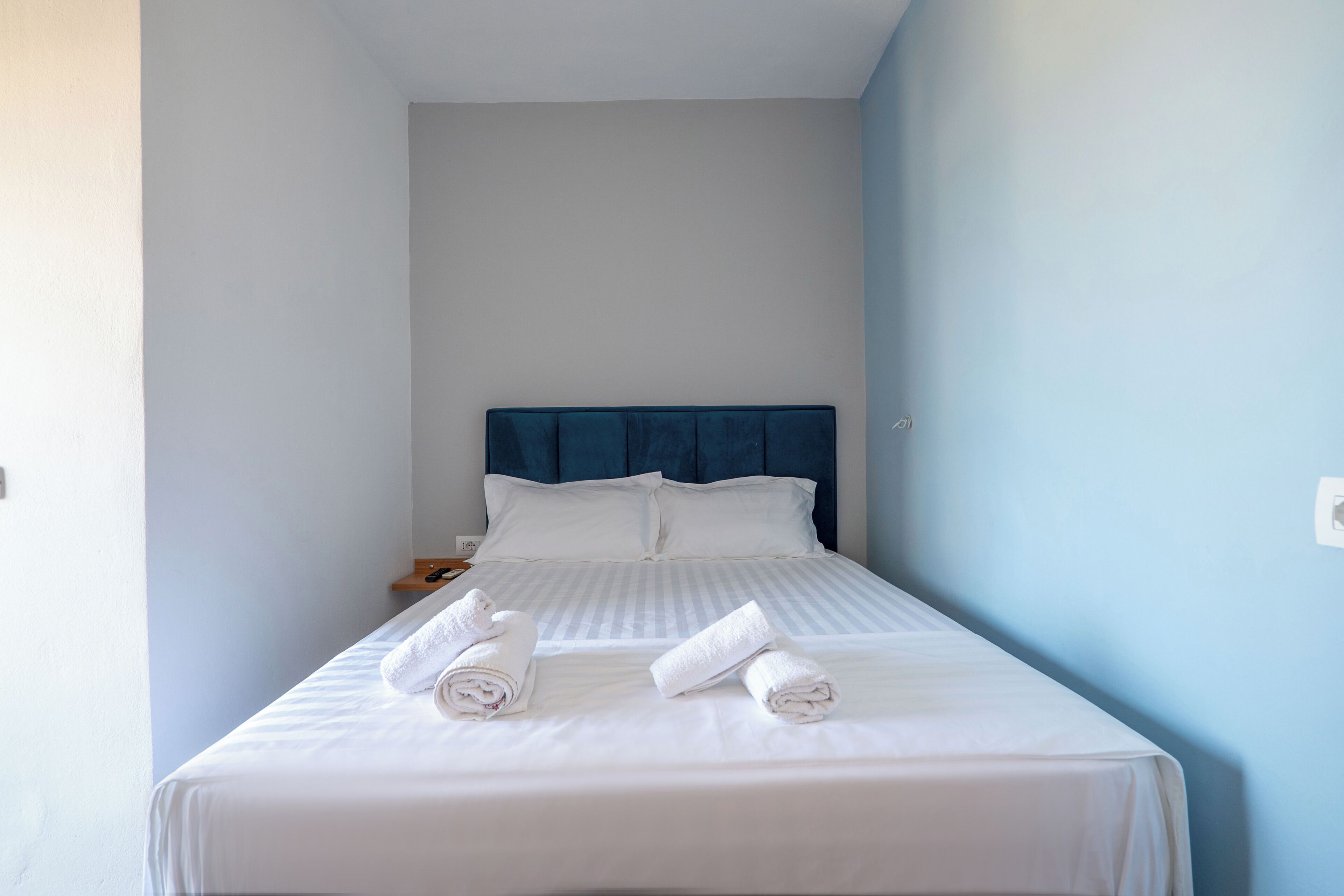 A single bed with white linens and two rolled towels, a blue headboard, and a nightstand with a phone.