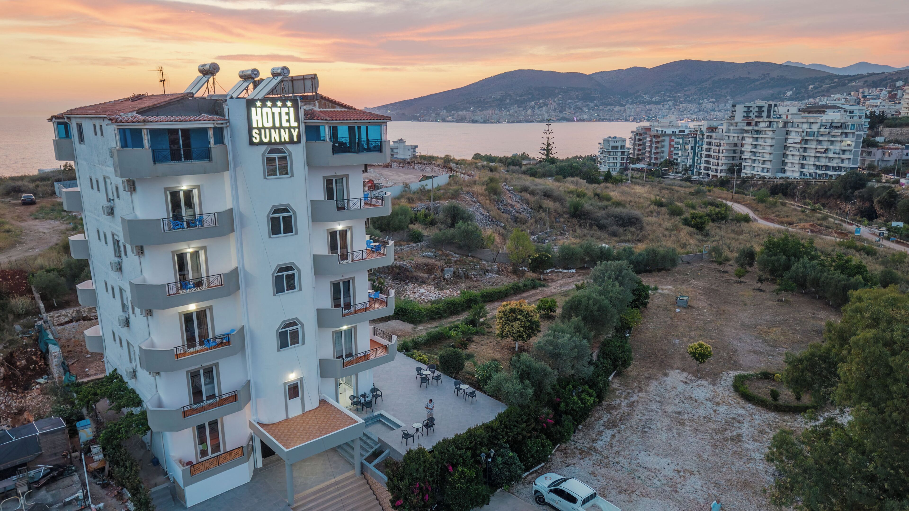 A multi-story hotel with balconies, a rooftop terrace, and a view of the sea and mountains.