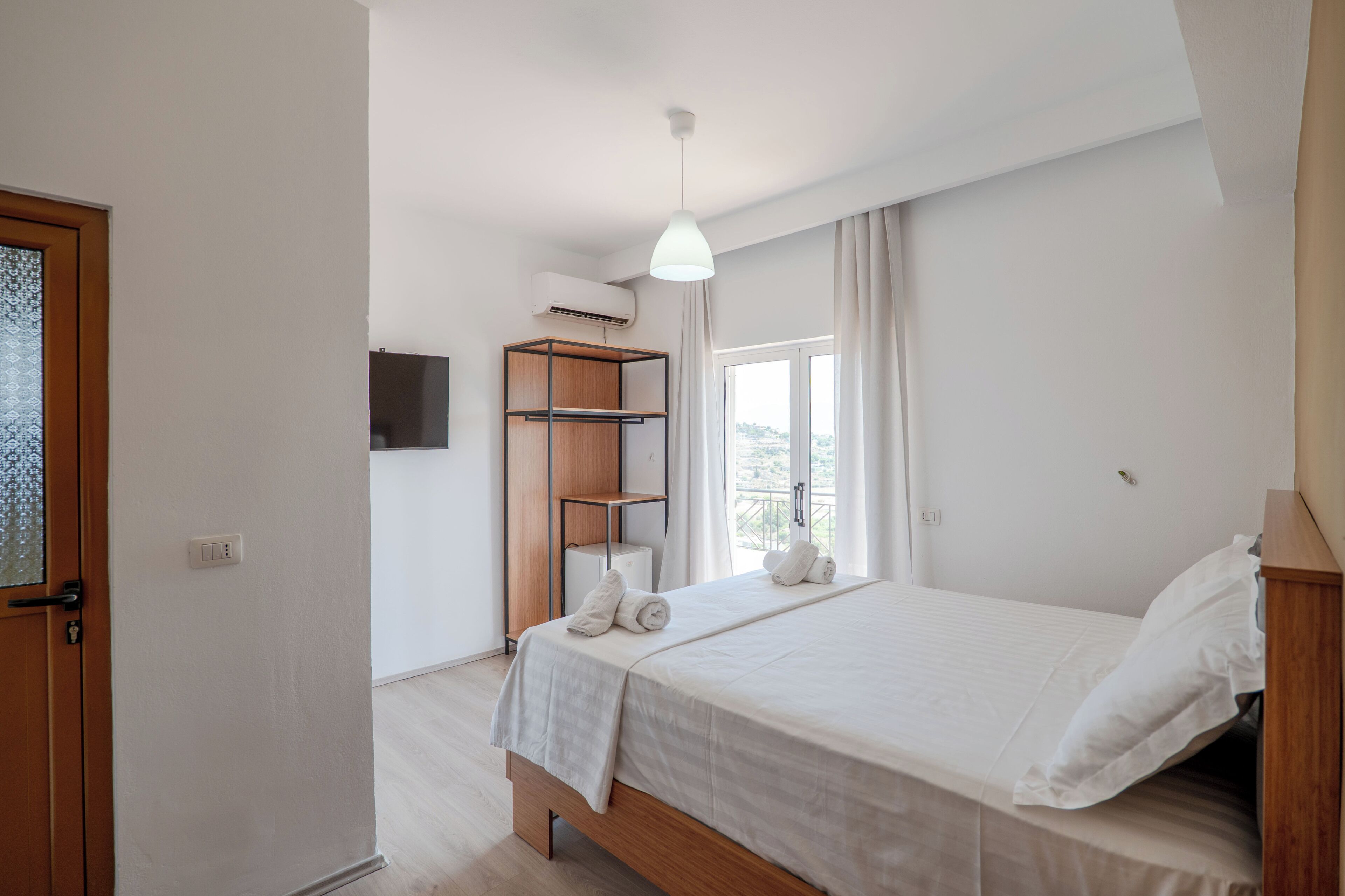 A bedroom with a bed, a wooden shelf unit, a TV, and a window with curtains.