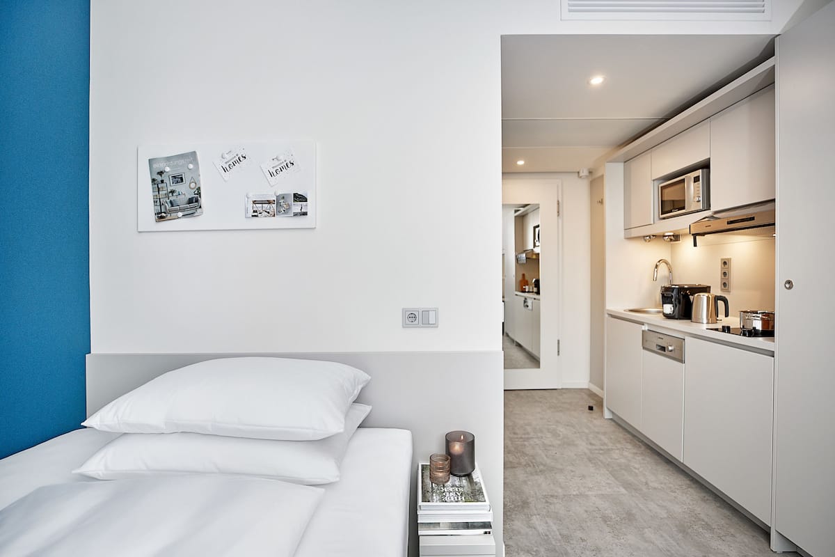 A modern hotel room with a neatly made bed, a small bedside table with a book and a candle, and a kitchenette with a microwave and coffee maker.