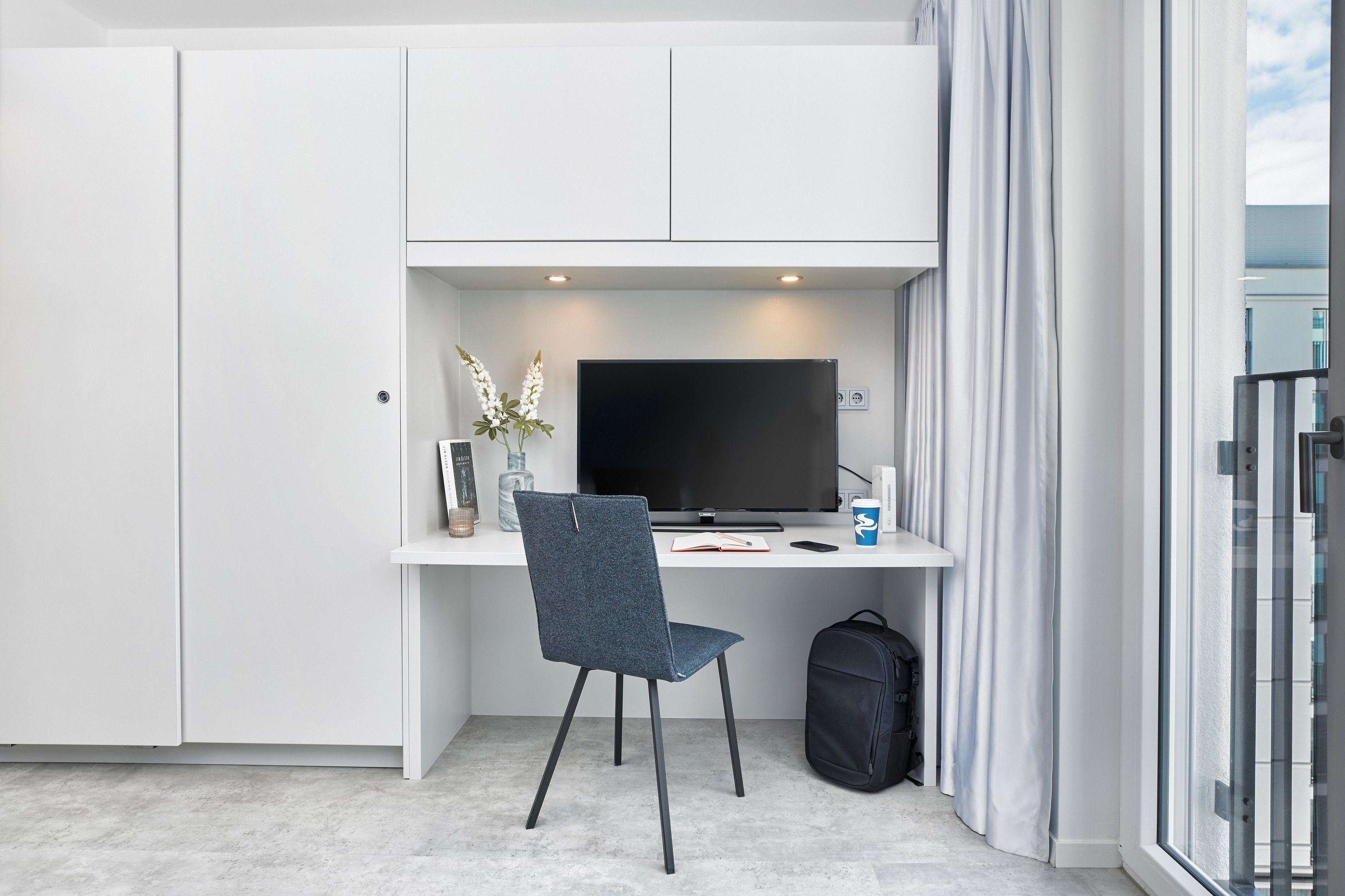 Une chambre moderne avec un bureau blanc, un téléviseur à écran plat, une chaise bleu foncé, un sac à dos noir et une fenêtre avec des rideaux.
