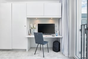 A modern room with a white desk, a flat-screen TV, a dark blue chair, a black backpack, and a window with curtains. - BD Munich Olympiapark (Munich)