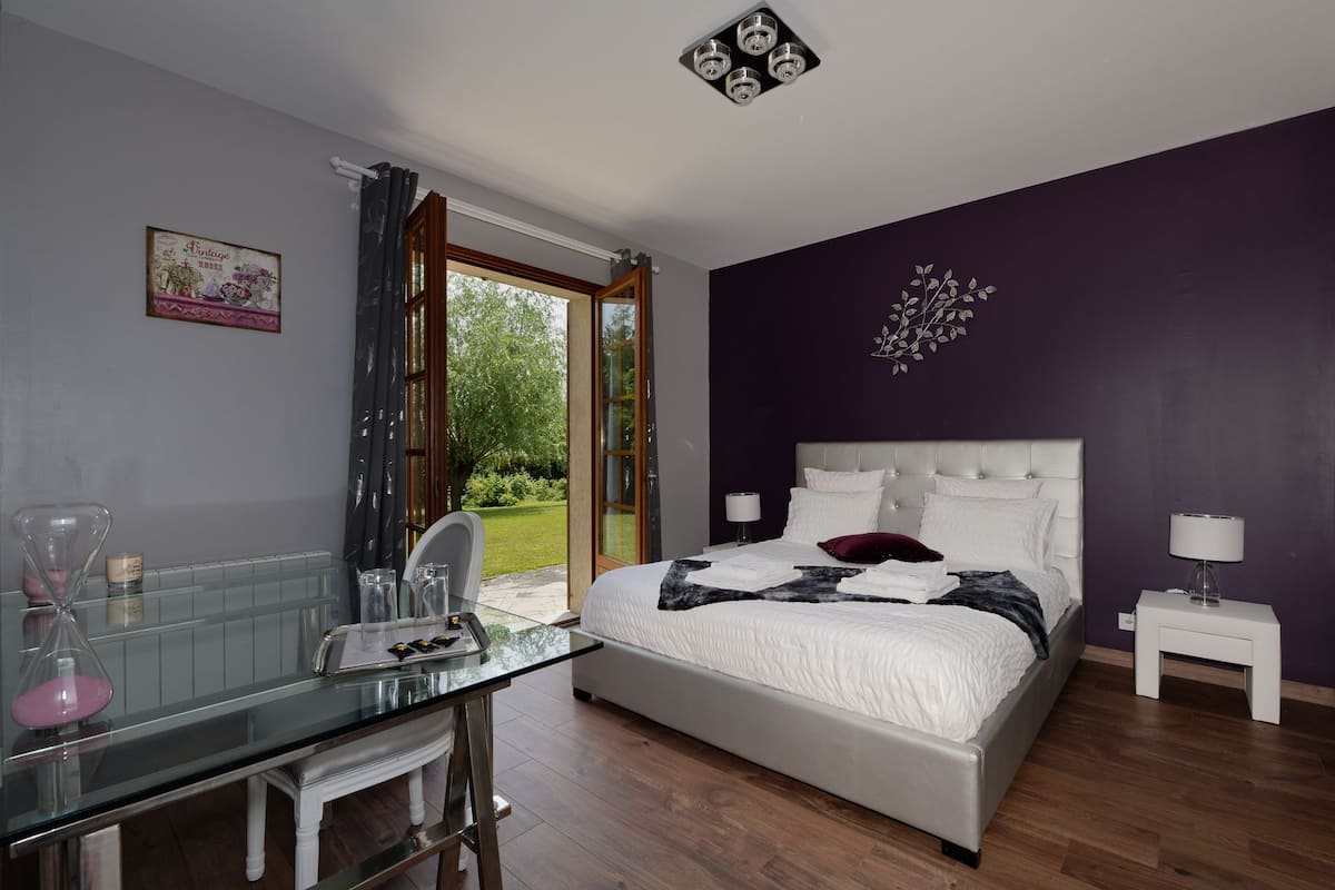 Une chambre à coucher avec un lit, des tables de chevet, une table en verre sur laquelle sont posés divers objets, une fenêtre avec des rideaux et une vue sur la verdure à l’extérieur.