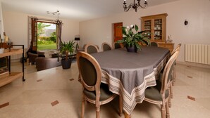 A dining room with a round table, six chairs, and a chandelier. There is a window with curtains, a cabinet with glassware, and a potted plant on the table. - Le Clos Magedon (Médan)
