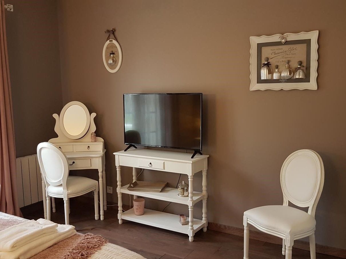 Une pièce avec un téléviseur posé sur un meuble blanc, deux chaises blanches, une coiffeuse avec un miroir et un certificat encadré accroché au mur.