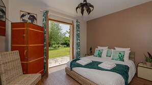 A bedroom with a large bed, a wicker chair, a wardrobe, and a view of the garden. - Le Clos Magedon (Médan)