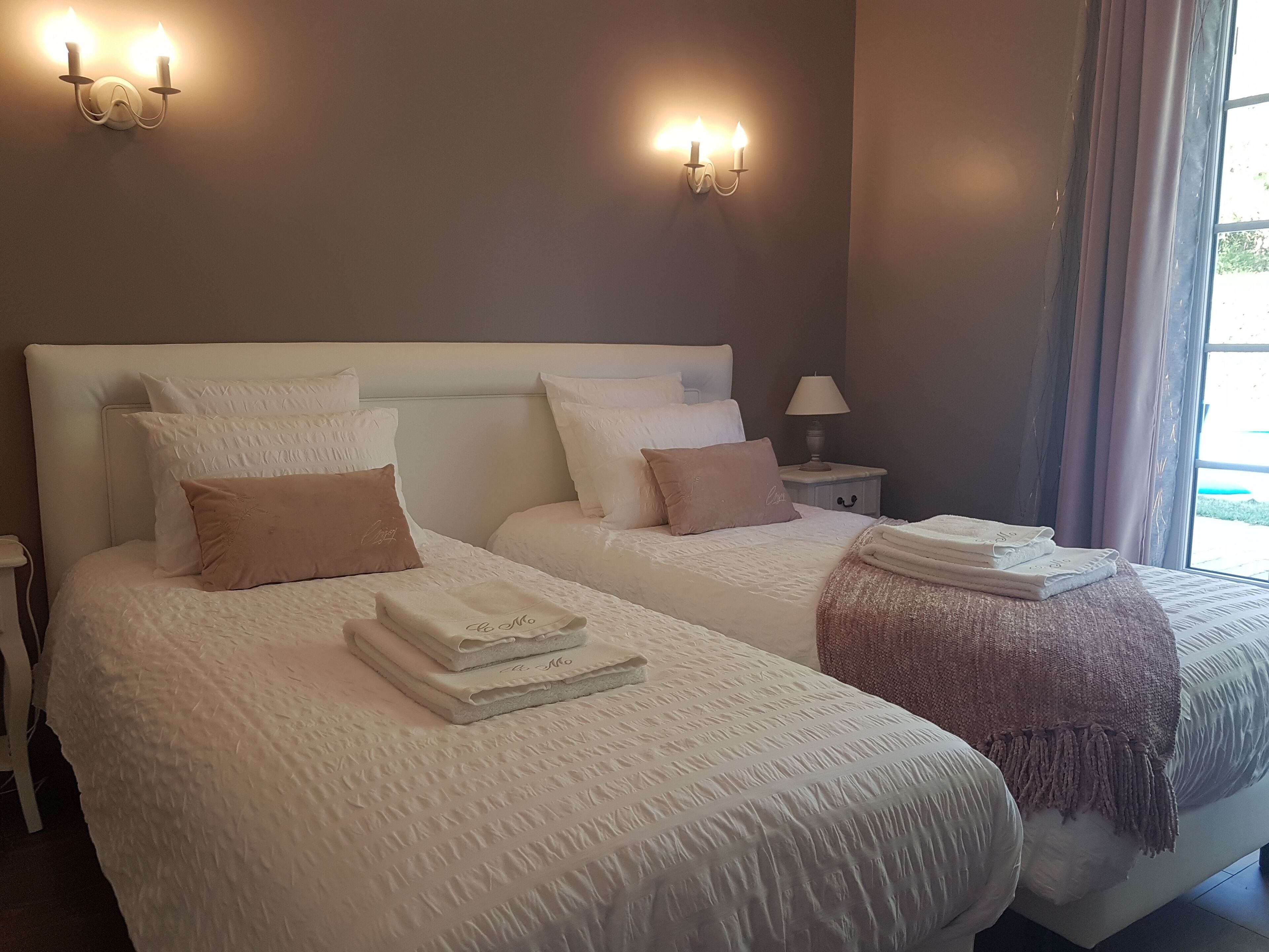 A hotel room with two beds, each with white bedding and a headboard. There are wall sconces with candles, a nightstand with a lamp, and a window with curtains.