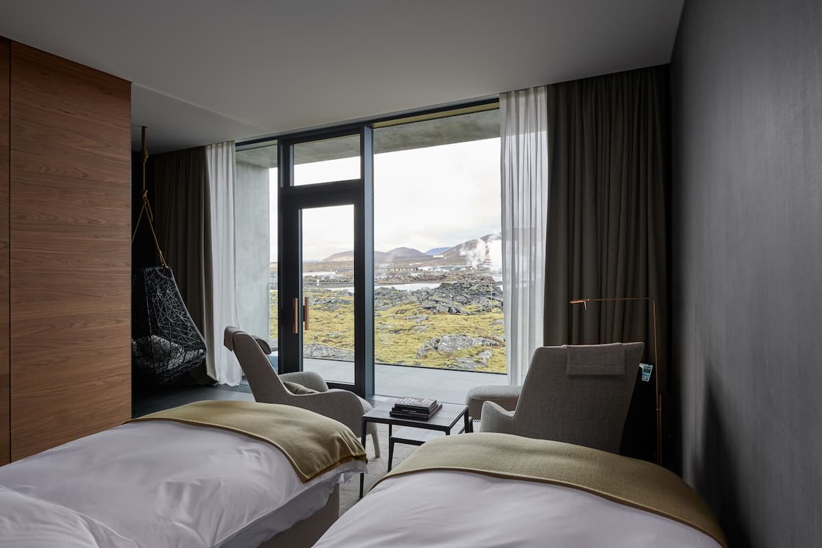 A modern hotel room with two beds, a hanging chair, and a view of a mountainous landscape.