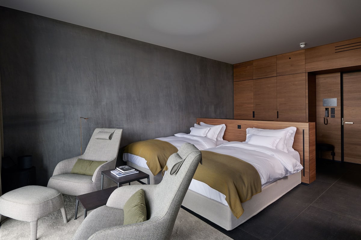 A modern hotel room with a large bed, two armchairs, a small table, and a wardrobe.