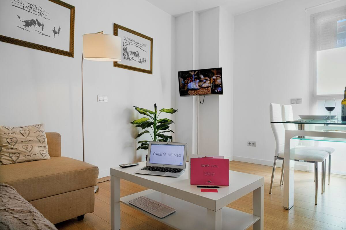 Una sala de estar moderna con un sofá, una mesa de centro con una computadora portátil, un televisor y un comedor con una mesa de vidrio y sillas.