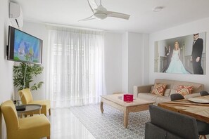 Ein modernes Wohnzimmer mit einer Couch, einem Couchtisch und Sesseln. Ein Fernseher ist an der Wand montiert, darüber hängt ein Gemälde.