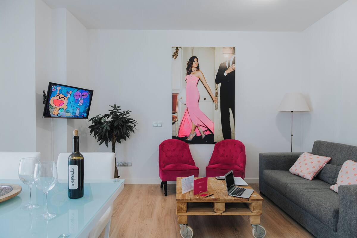 Una sala de estar moderna con un televisor de pantalla plana, una botella de vino, dos sillones rojos, un sofá gris y una mesa de centro de madera.