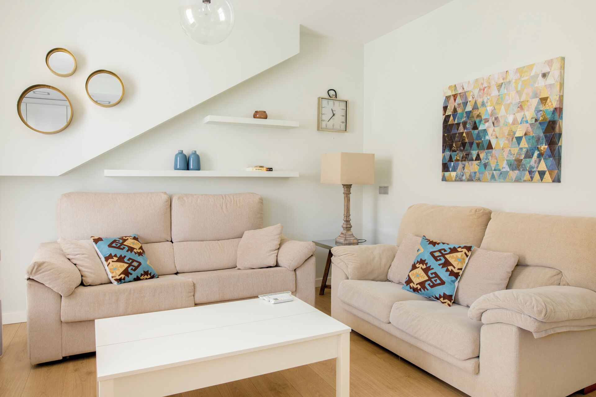 A living room with two beige sofas, a white coffee table, a wall clock, a lamp, and a geometric art piece.