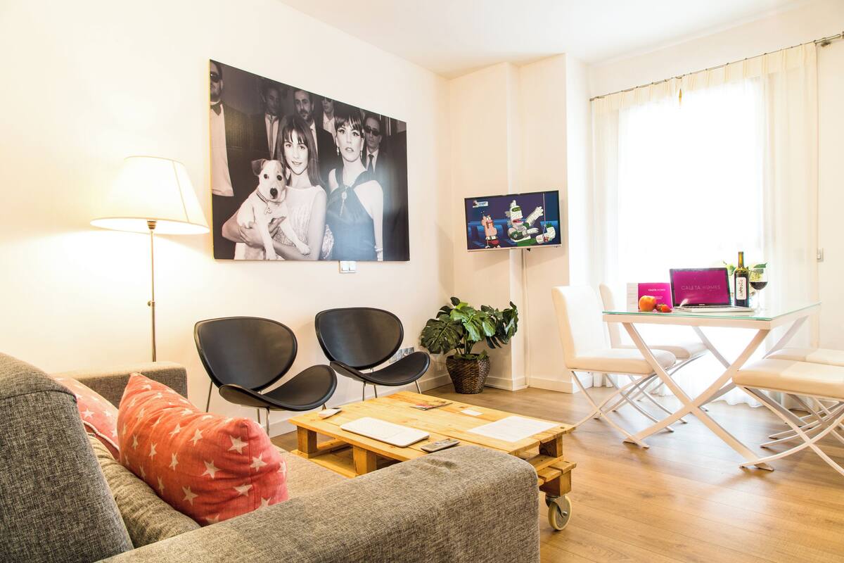 Una sala de estar con un sofá, dos sillas negras, una mesa de centro de madera, una lámpara, un televisor, una mesa de comedor con sillas y una planta en maceta.