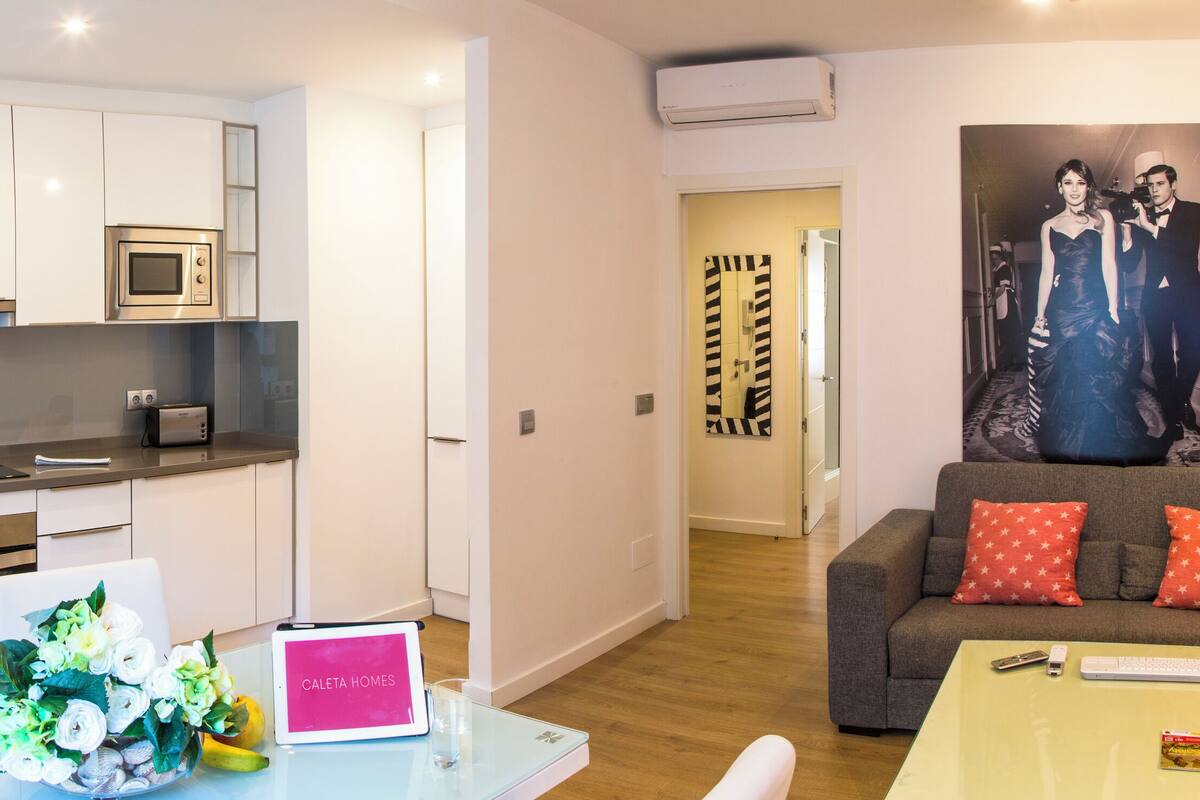 Una sala moderna con un sofá gris, una mesita y un comedor con una mesa blanca y flores. Se observa una cocina con armarios blancos y un microondas.