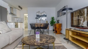 Un salon moderne avec un canapé, une table basse ronde, un coin repas avec des chaises, un téléviseur et une cuisine en arrière-plan.