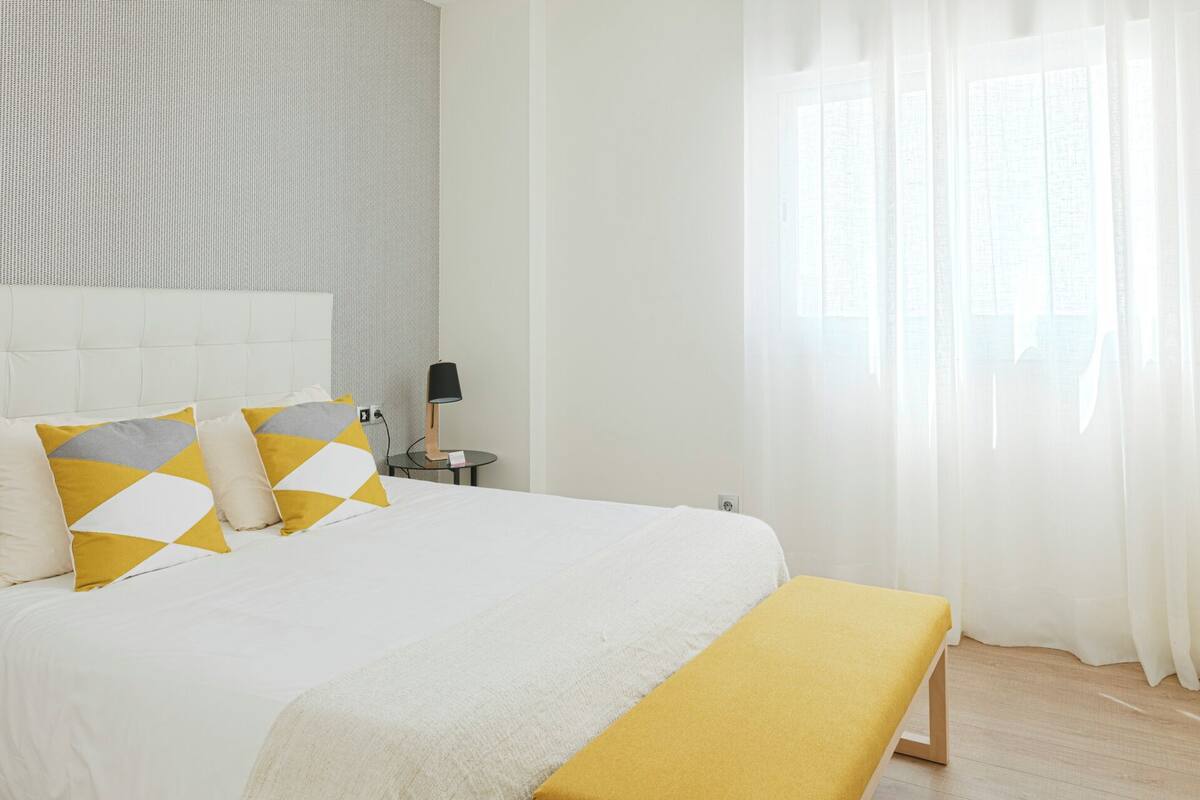 Habitación de hotel impecable con una cama grande, una mesita de noche con lámpara y cortinas translúcidas que dejan entrar luz natural.
