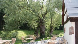An outdoor patio with wooden tables and chairs, surrounded by lush greenery and a large tree. - Landhaus Hui Wäller (Greifenstein)