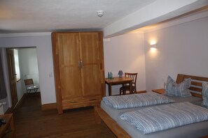 Un dormitorio con un armario de madera, un escritorio con silla, dos camas con ropa de cama a rayas y una mesita de noche con lámpara.