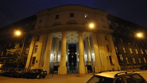 Ein großes, historisches Gebäude mit einem prunkvollen Eingang, das eine zentrale Arkade und Säulen aufweist und von parkenden Autos umgeben ist.