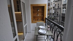 Ein Balkon mit Tisch für zwei Personen, ein Holzregal mit Dekoration und Blick auf benachbarte Gebäude.
