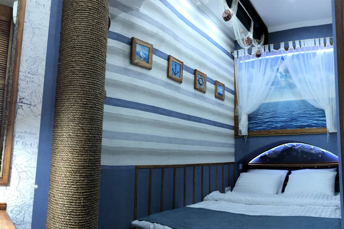 A bedroom with a striped wall, a large window with a view of the sea, a bed with white bedding, and a rope cat tree.