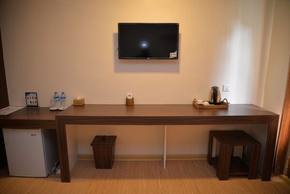Una habitación con un escritorio de madera grande, un televisor montado en la pared, un refrigerador pequeño y un cesto de basura.