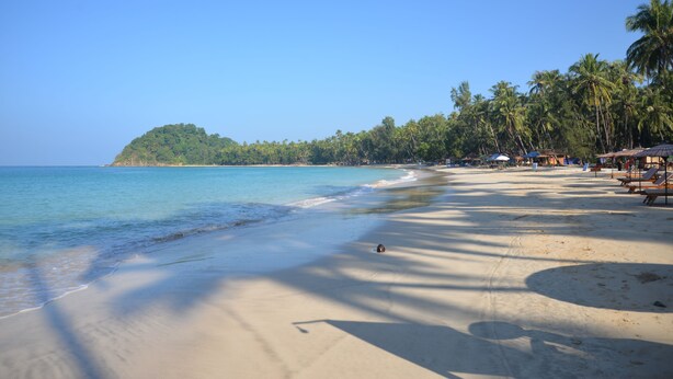 A beach with clear blue water, sandy shore, palm trees, and beach umbrellas. - Thazin Palace A Hotel (Ngapali)