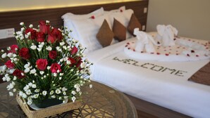 A neatly made bed with a "WELCOME" sign, a basket of red and white flowers, and a small table with a geometric patterned glass top. - Thazin Palace A Hotel (Ngapali)