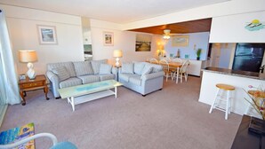 A living room with a sofa, two chairs, a coffee table, a dining table with chairs, a kitchen area with a refrigerator, and a ceiling fan. - Beachmark 3F - 1 Bedroom Condo by RedAwning (Ocean City)