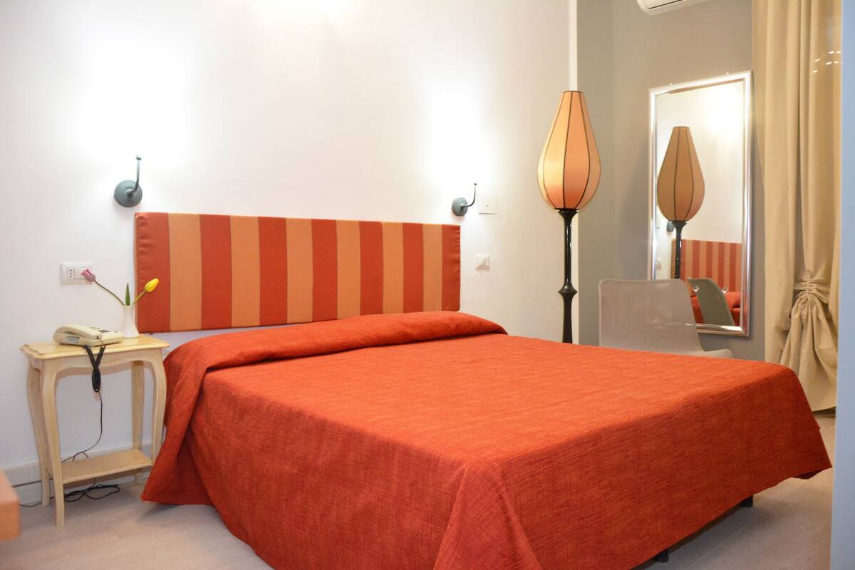 A hotel room with a bed, a bedside table, a lamp, and a mirror.
