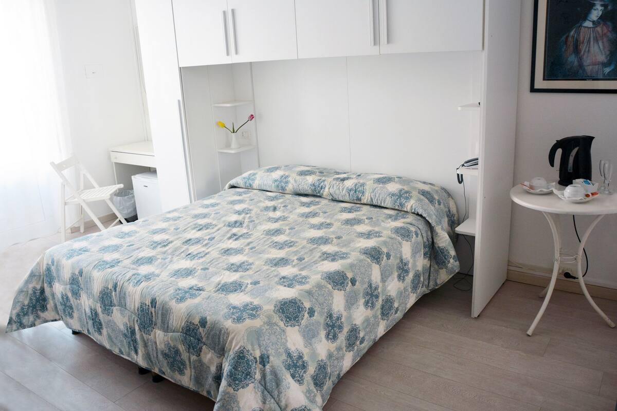 A bedroom with a bed, a desk, and a small table with a kettle and cups.