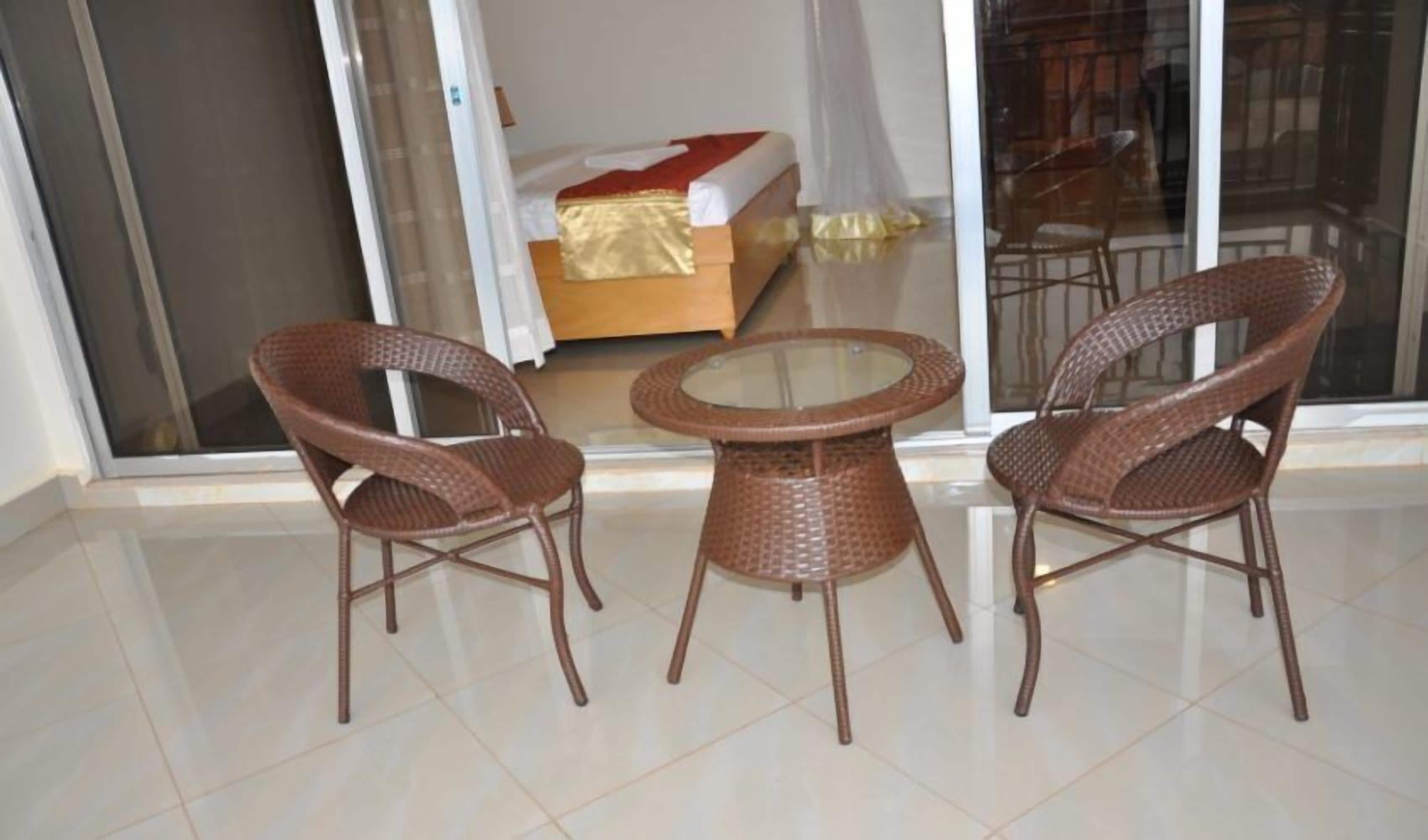A room with a glass door leading to a balcony, a bed with a red and white bedspread, and two wicker chairs around a small glass table.