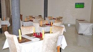A dining area with tables set for four, each with a white tablecloth, candles, and red napkins. - EKA Hoima Hotel (Hoima)