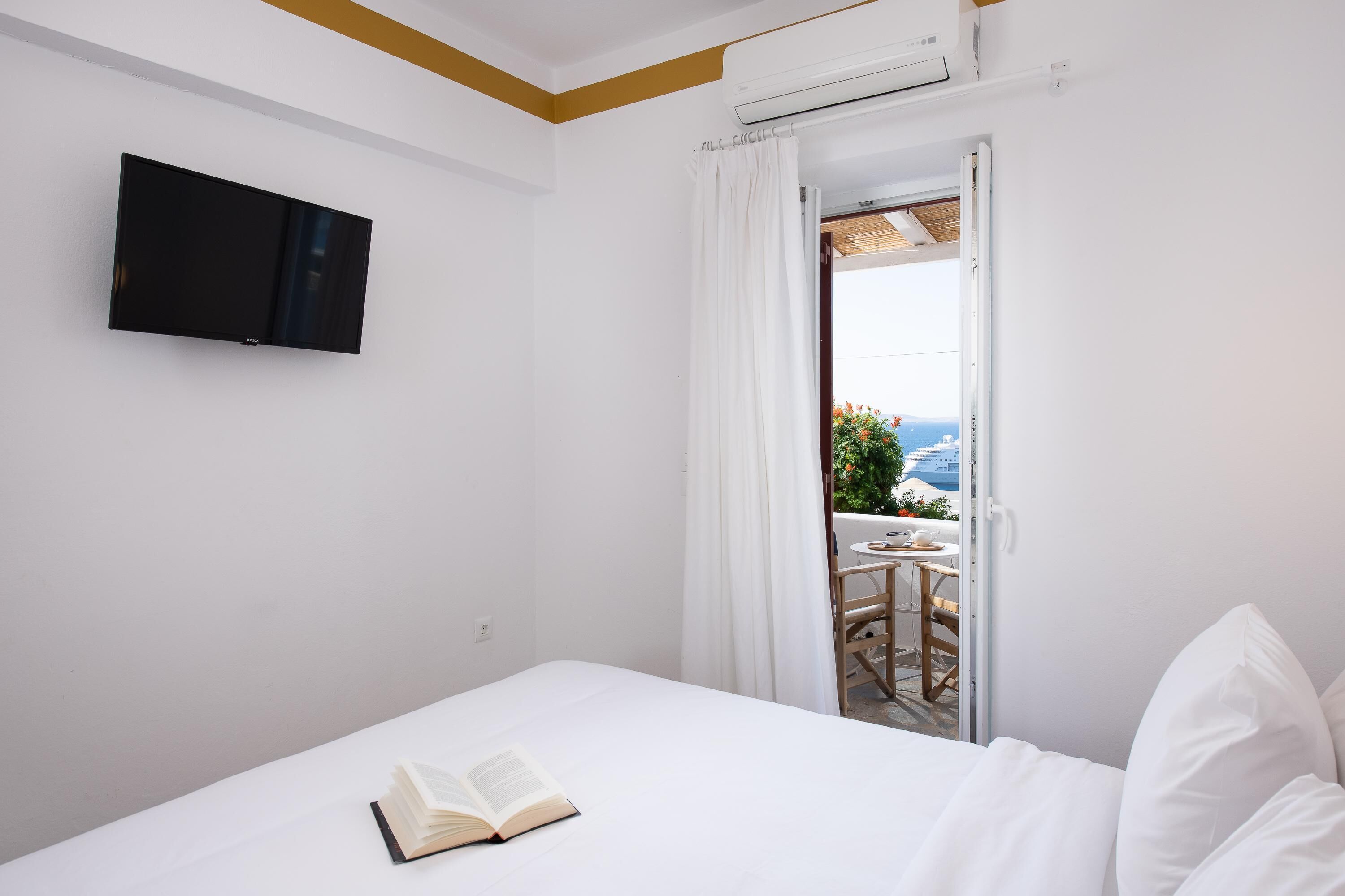 Une chambre d’hôtel avec un lit, un livre ouvert, un téléviseur fixé au mur et un balcon avec une table et des chaises.