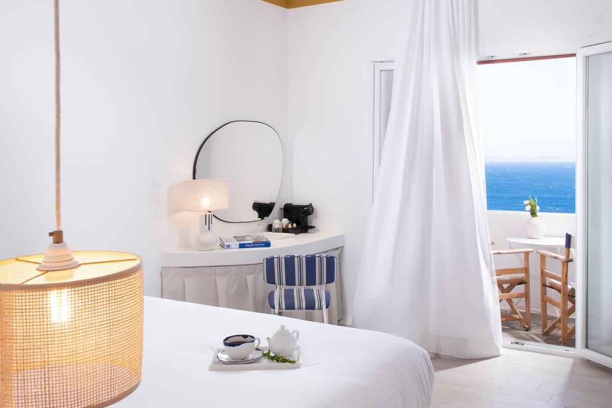 Habitación de hotel con cama, mesita de noche con lámpara, espejo, ventana con vista al mar y balcón con mesa y sillas.