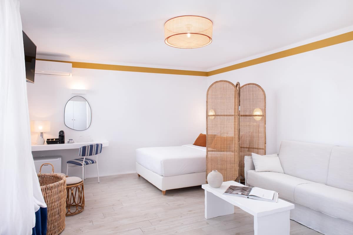 Una habitación de hotel moderna con una cama blanca, un sofá blanco, una mesita de centro blanca, una silla de mimbre, una cesta de mimbre, un espejo y una lámpara en el techo.