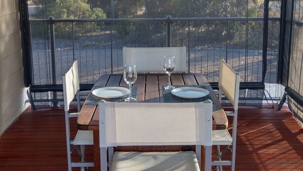 An outdoor dining area with a table set for two, chairs, and a view of trees through a screened balcony.