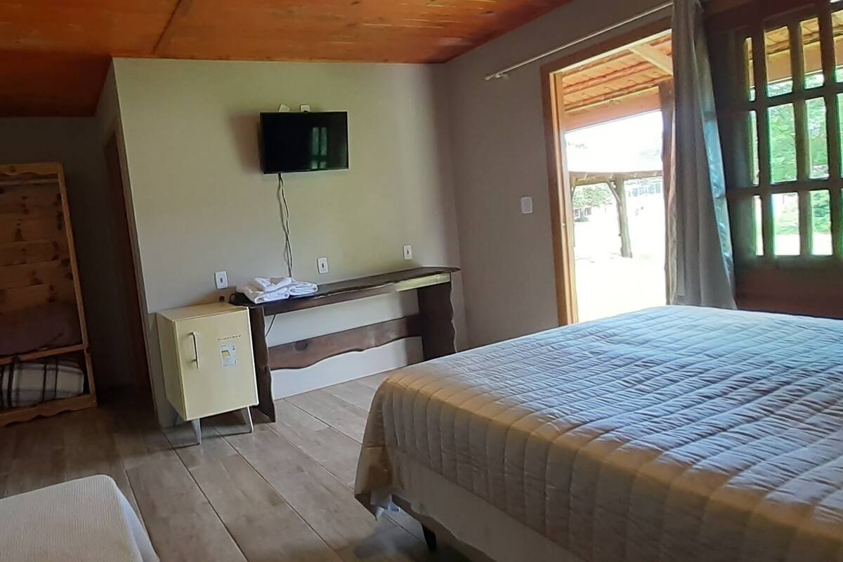 Quarto com cama, guarda-roupa de madeira, pequena escrivaninha com televisão e janela com vista para vegetação.