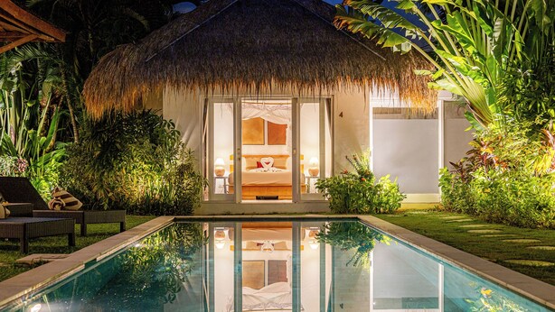 Un bungalow au toit de chaume, doté d’une piscine et entouré d’une végétation luxuriante.