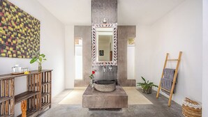 Ein Badezimmer mit einem Steinkonstruktion-Waschbecken, einem Spiegel, einem Holzbodenregal mit einer Vase und einer Pflanze, sowie einer an der Wand stehenden Leiter.